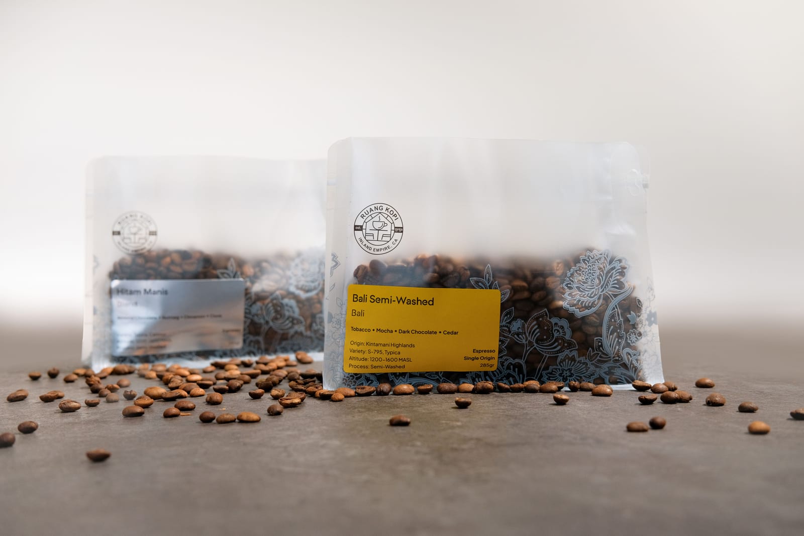 Two Ruang Kopi bags, the Hitam Manis blend and the Bali semi-washed, sitting on loose roasted beans.