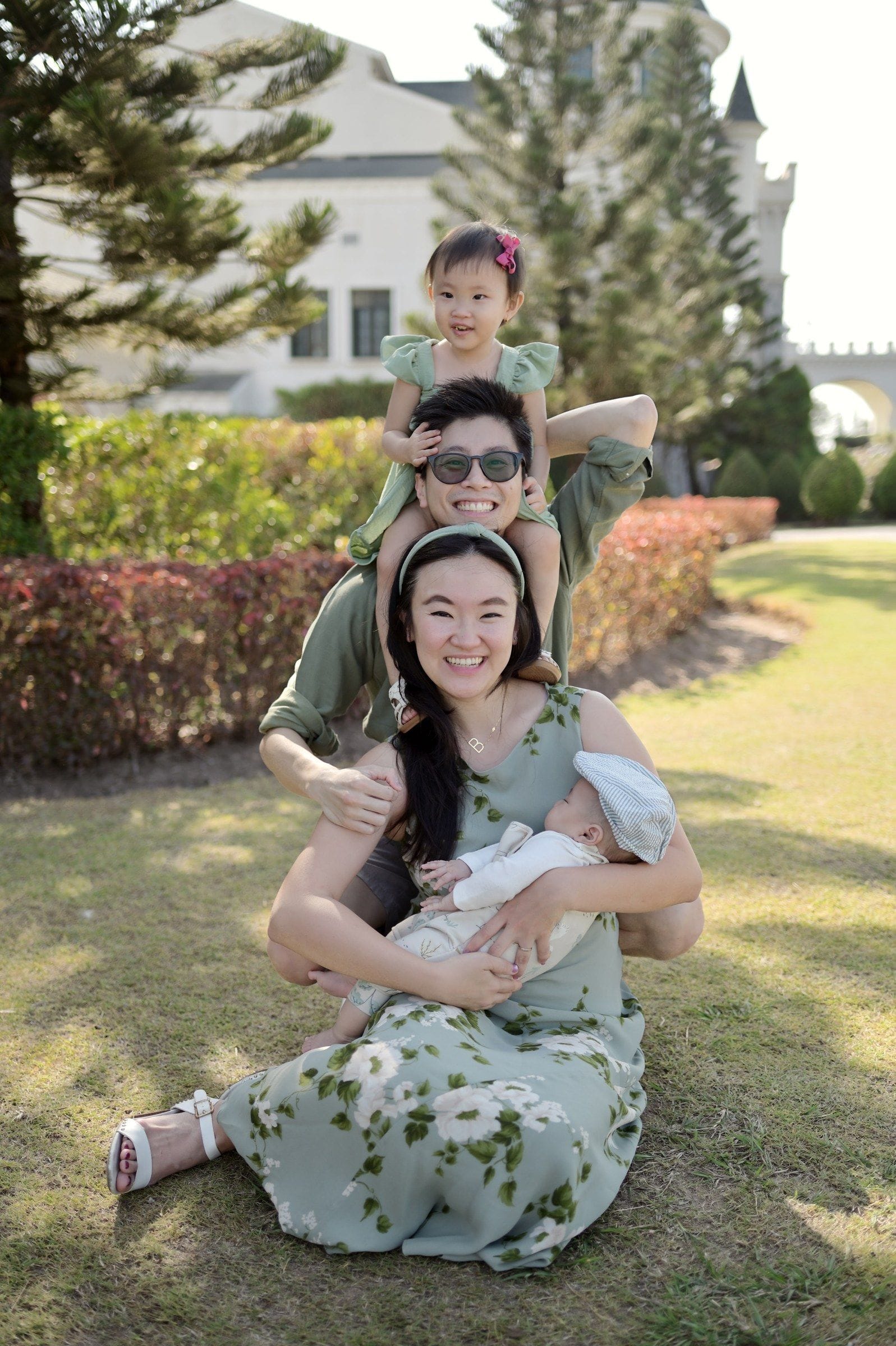 Rei and Bella outdoors with their toddler on Rei's shoulders and their newborn in Bella's arms.