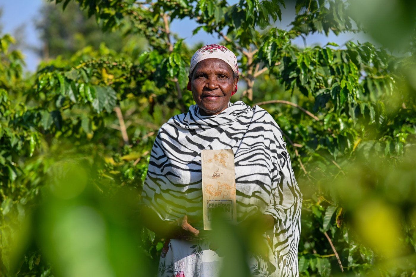 Margaret Kuria, farmer and co-owner of Liwani Coffee Estate, standing among coffee trees in Solai holding a bag of Solai Coffee Precious Bean.