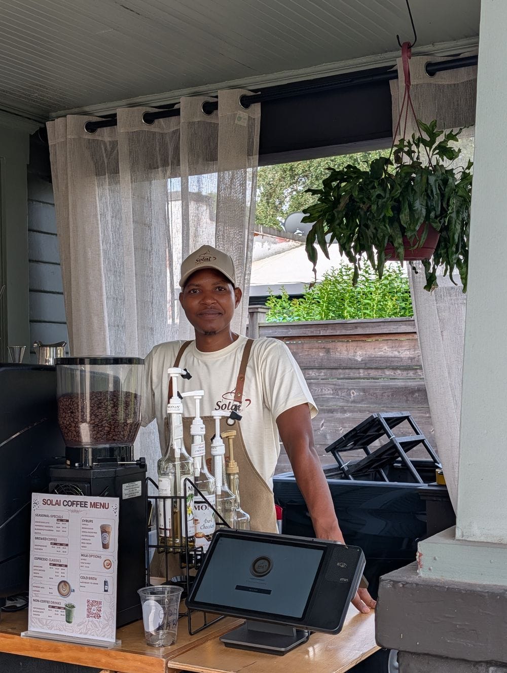 Peter Kuria behind the counter of the Solai Coffee stand in Orlando, in a Solai cap and apron, with the menu board and a grinder in front of him.