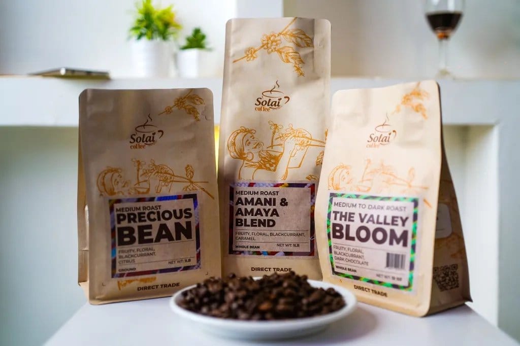 Three Solai Coffee bags on a white counter, Precious Bean, Amani and Amaya Blend and The Valley Bloom, with a plate of roasted beans in front.